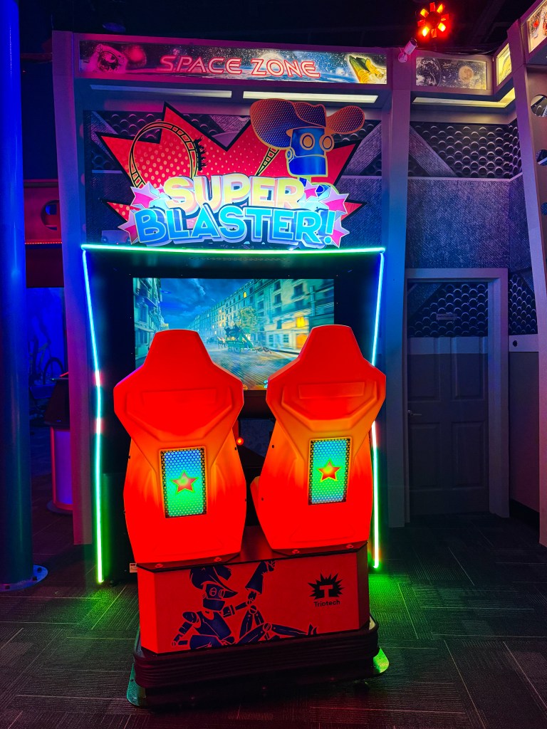 A brightly lit arcade game titled 'Super Blaster!' featuring two red gaming seats, set against a colorful backdrop in a space-themed environment.