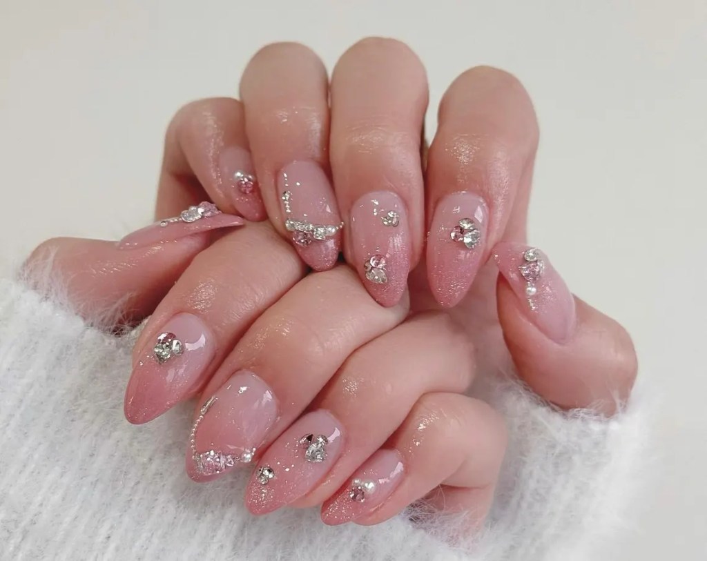 A close-up of hands with manicured nails featuring a glossy nude base, silver glitter, and rhinestone embellishments.