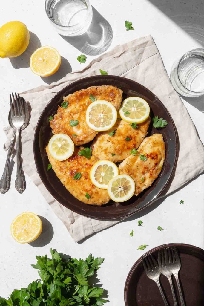 A plate of golden-brown chicken breasts garnished with lemon slices and parsley, accompanied by a slice of lemon and fresh parsley on the side.