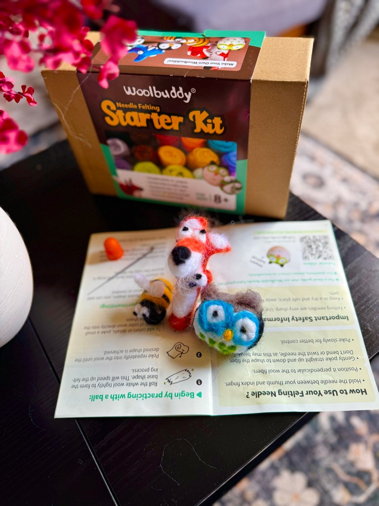 A Woolbuddy Needle Felting Starter Kit box displayed on a table, surrounded by colorful felted animal figures including a fox, owl, and bee, along with an instruction booklet.