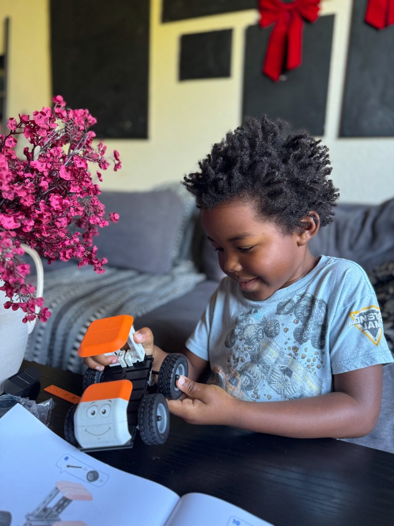 A young boy with curly hair intently assembles a small toy vehicle while sitting at a table with a pink flower arrangement and an open instruction booklet.