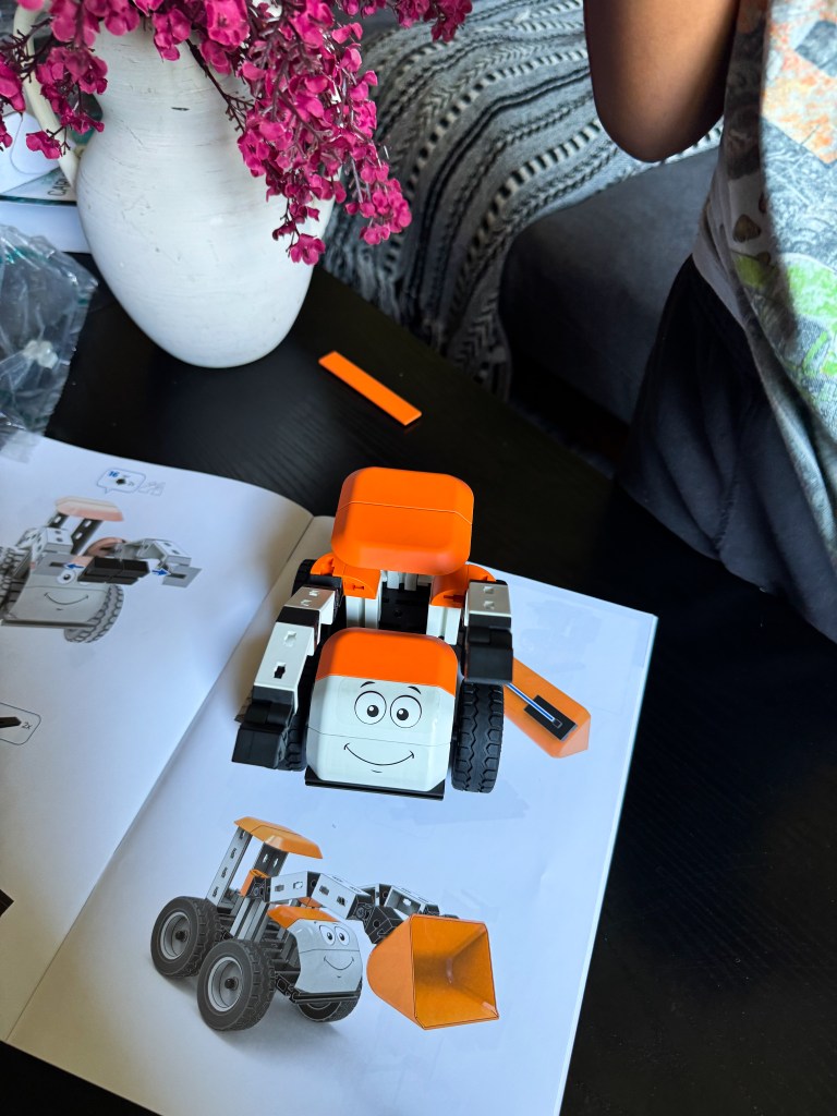 A colorful toy vehicle with a smiling face, partially assembled, placed on an instruction manual featuring an illustration of the completed model. A vase with pink flowers and a few scattered toy parts are visible in the background.