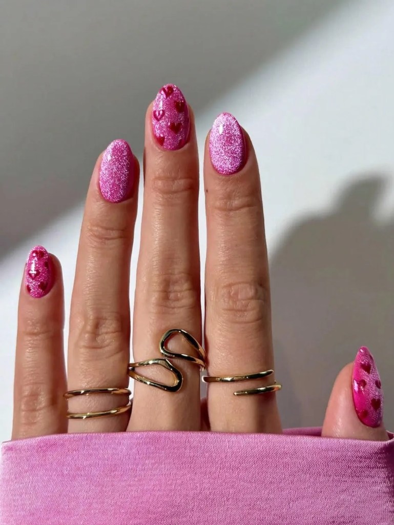A close-up of a hand with manicured nails featuring pink glitter and heart designs, adorned with several gold rings, against a light background.