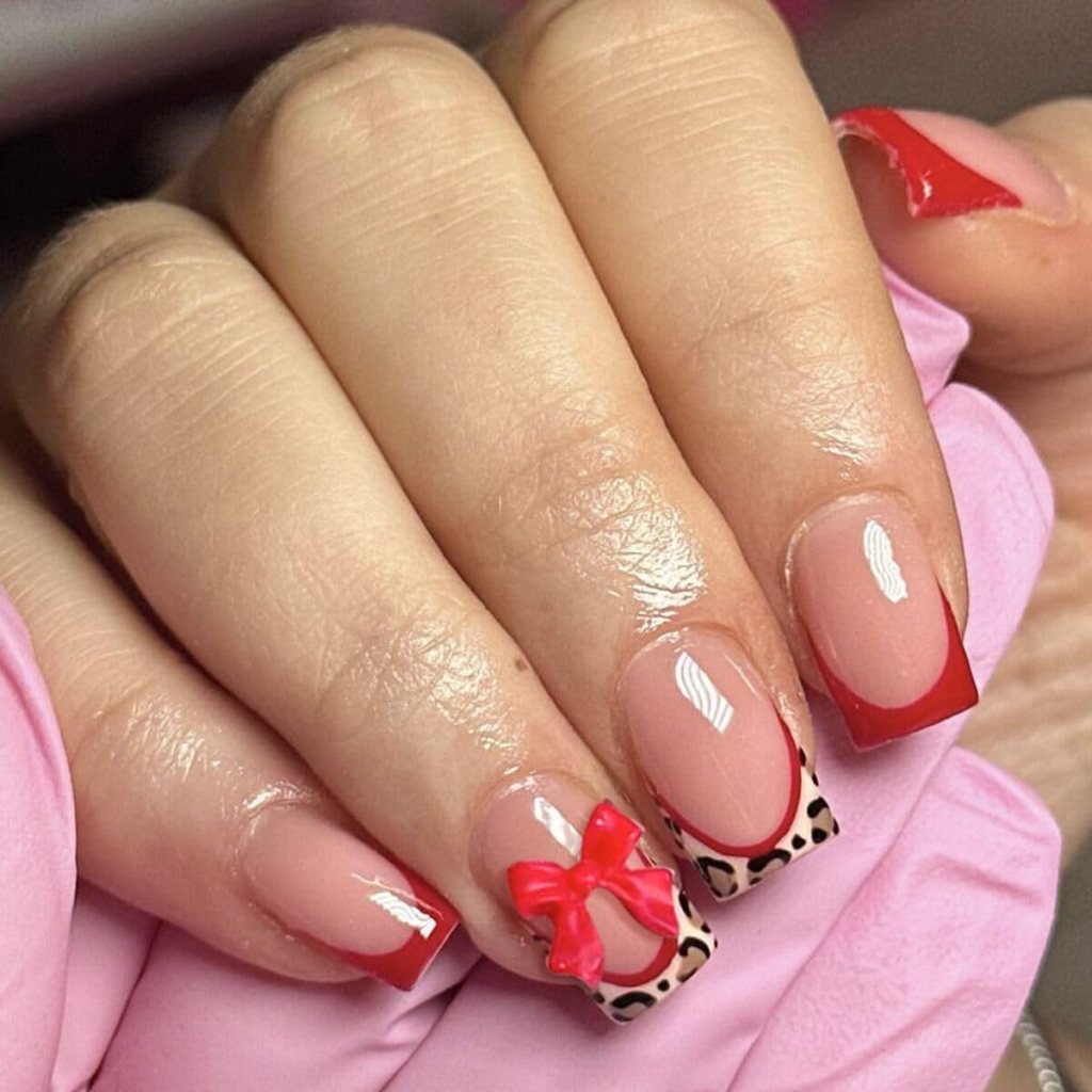 Close-up of a hand with manicured nails featuring a mix of nude polish, red tips, and a leopard print accent nail with a red bow.