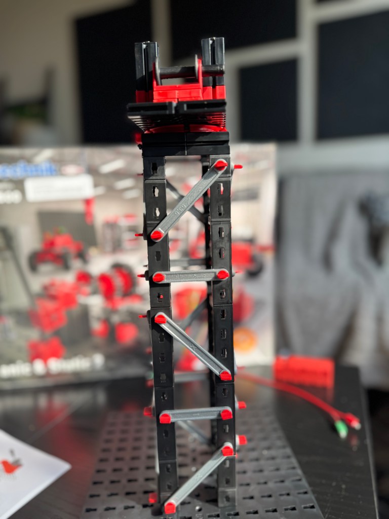 A completed model from the Fischer Technik STEM Kit, showcasing a tall structure made of black and red building pieces, set against the backdrop of its packaging.