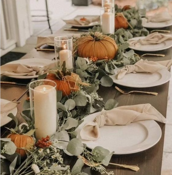 A cozy fall dining table decorated with orange pumpkins, eucalyptus foliage, and white candles, featuring neutral napkins on each plate.