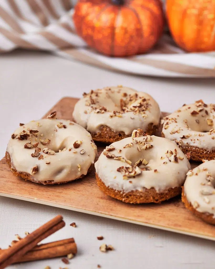 A wooden platter holding glazed pumpkin spice donuts topped with chopped nuts, with decorative pumpkins in the background.