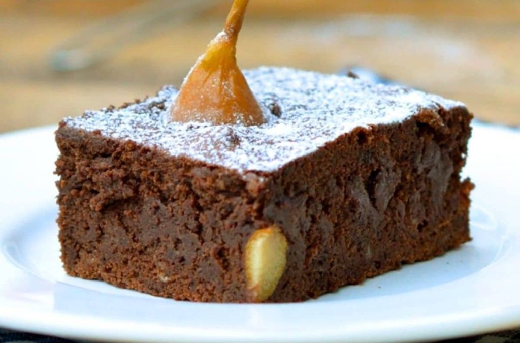 A slice of rich chocolate brownie topped with a pear slice and dusted with powdered sugar, served on a white plate.