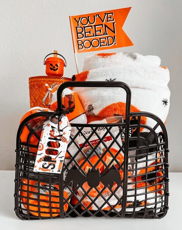 A black basket filled with orange-themed Halloween goodies, including a cozy blanket, treats, and a flag that says 'YOU'VE BEEN BOOED!'