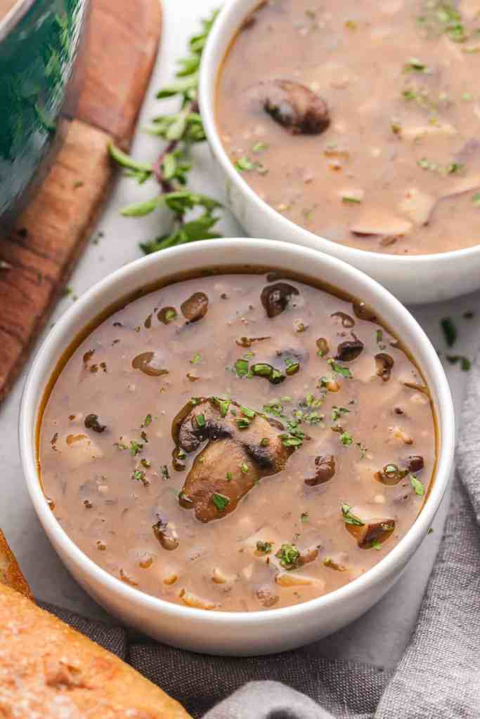 A warm bowl of dairy-free cream of mushroom soup garnished with fresh herbs, served alongside a slice of bread.