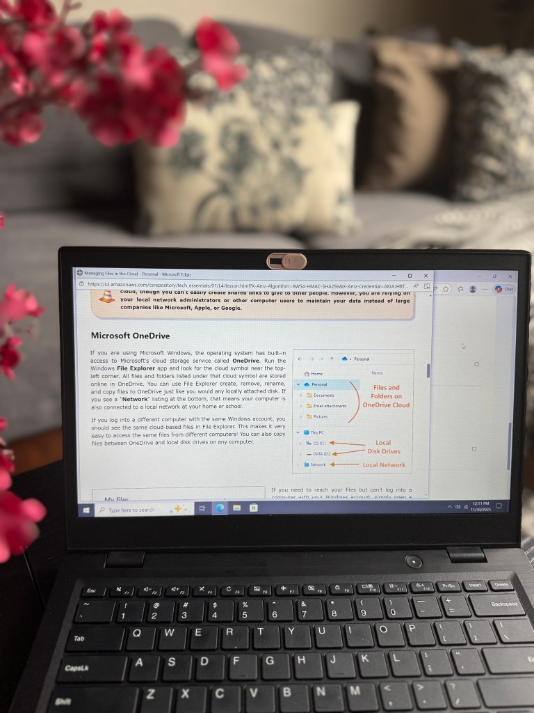 A laptop displaying a OneDrive tutorial on managing files in the cloud, with a floral background and a soft-focused couch.