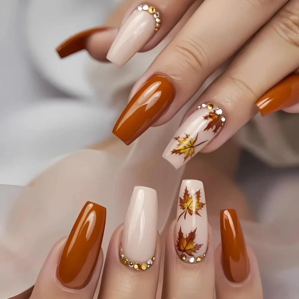 A close-up of manicured nails featuring a mix of burnt orange and nude polish, adorned with hand-painted fall leaves and rhinestone accents for a festive Thanksgiving look.