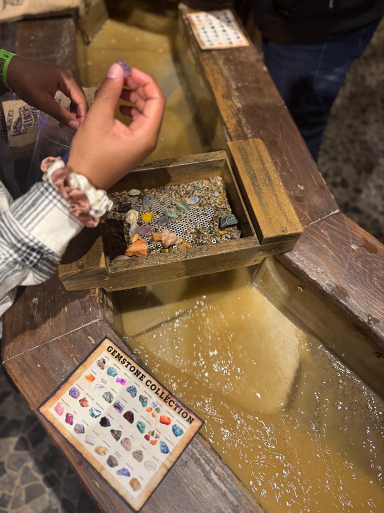 A child holding a gemstone after sifting through a water sluice at Great Wolf Lodge's Oliver's Mining Co., with a collection of colorful stones visible in the water.