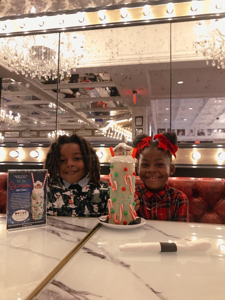 Two children smiling beside Sugar Factory Biloxi's festive Christmas milkshake topped with a miniature gingerbread house.