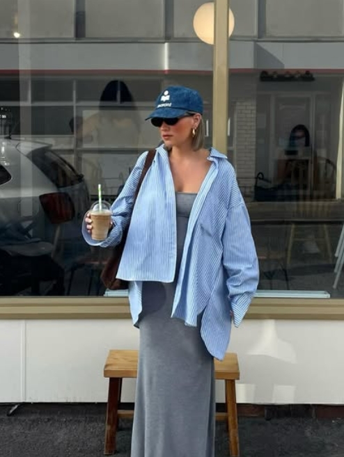 A woman standing outside a café, wearing a striped oversized shirt over a gray maxi dress, holding a drink, with a denim cap and sunglasses.