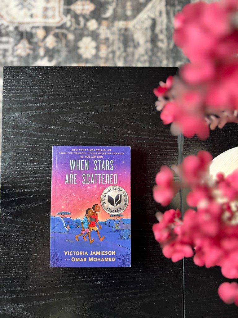 A close-up view of the graphic novel 'When Stars Are Scattered' by Victoria Jamieson and Omar Mohamed, resting on a dark wooden table, with a floral arrangement in the foreground.
