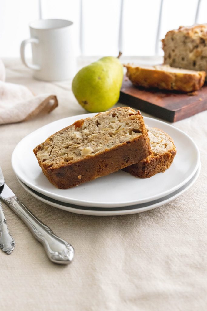 Slices of banana pear bread on white plates with a whole pear and a cup in the background.