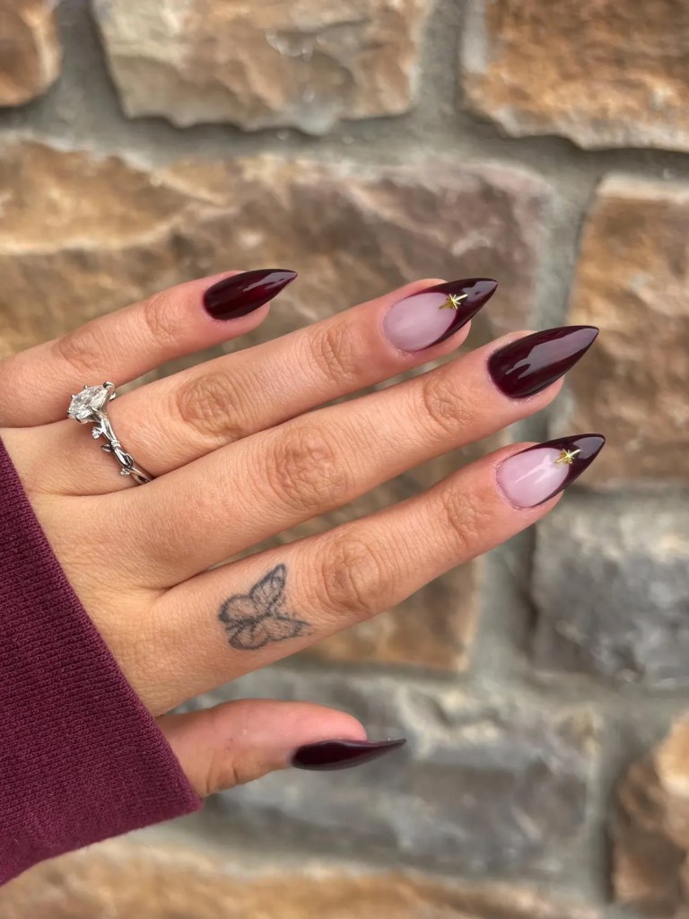 A close-up of a hand with long, pointed nails painted in a deep red color with one nail featuring a light pink design and a small decorative star. The hand is adorned with a silver ring and has a butterfly tattoo on the wrist. The background consists of a textured stone wall.