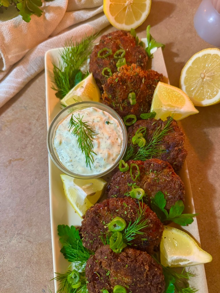 A platter of crispy golden-brown salmon patties garnished with fresh herbs, accompanied by a bowl of creamy dipping sauce and lemon wedges.
