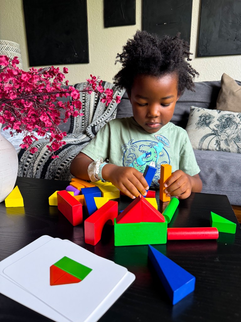 A young child playing with colorful geometric blocks on a black table, focusing on building structures. A guide card with shapes is visible nearby, and there are pink flowers in a vase in the background.