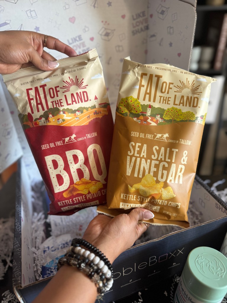 A hand holding two bags of 'Fat of the Land' kettle-style potato chips, one flavor labeled 'BBQ' and the other 'Sea Salt & Vinegar', displayed in a box with shredded paper filling.