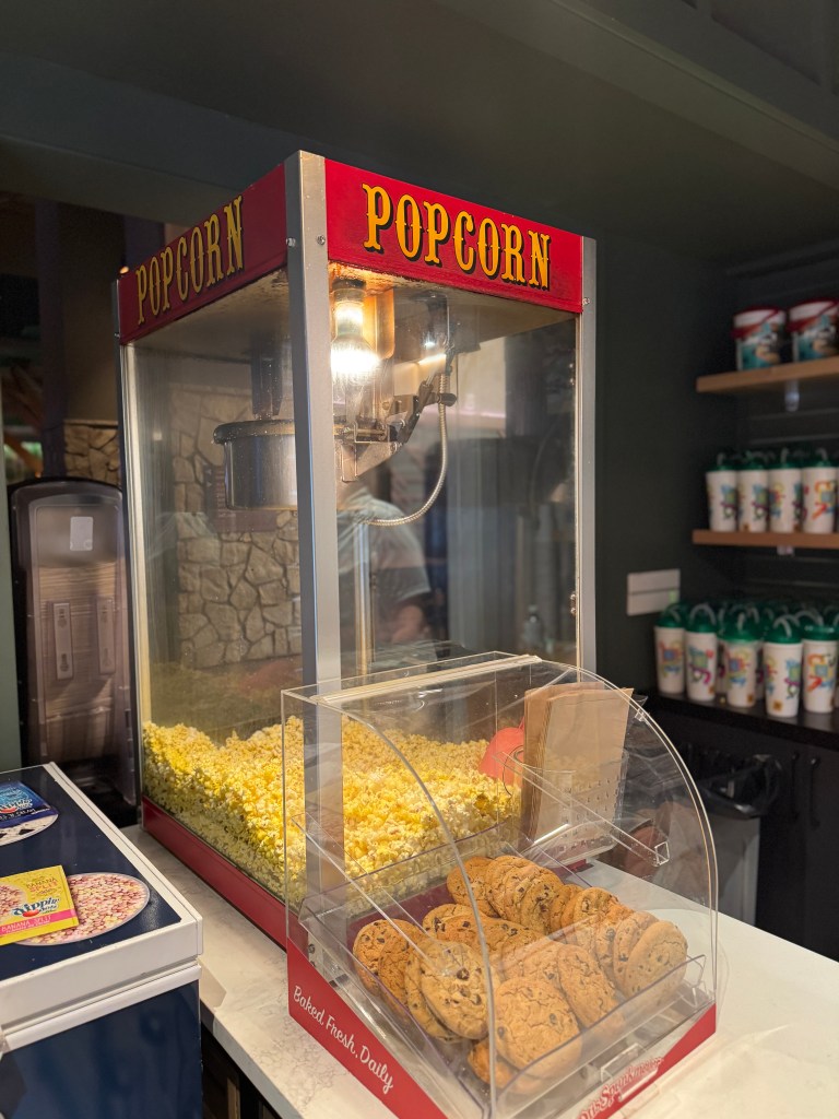 A popcorn machine with freshly popped popcorn and a display of baked cookies in a glass container, set against a backdrop of shelves filled with beverage cups. Great Wolf Lodge lobby area in Grapevine, TX 