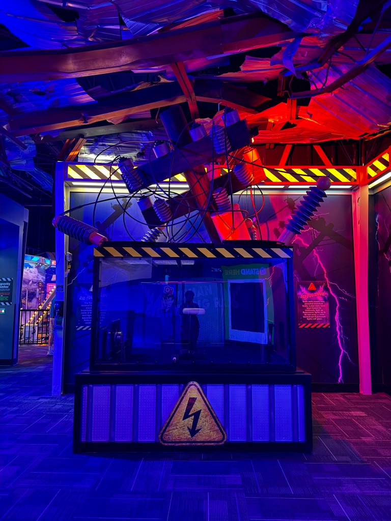 An exhibit featuring a glass container with a warning sign and electric cables, illuminated with blue and red lights, resembling a scientific demonstration of electricity.