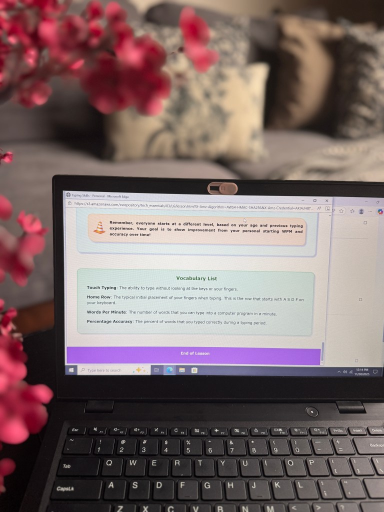 A laptop screen displaying a lesson on touch typing skills with a vocabulary list, including terms like 'Home Row' and 'Words Per Minute', framed by a flower arrangement in the foreground.