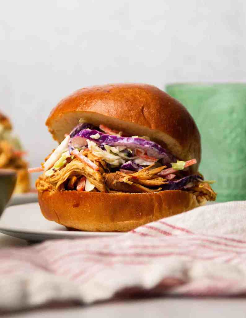 A close-up of a pulled chicken sandwich topped with coleslaw, served on a soft bun, with a blurred background.