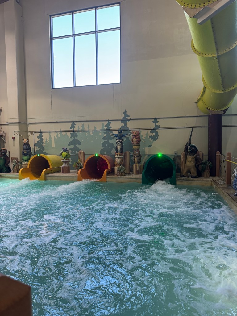Indoor water park featuring colorful water slides and a wave pool, surrounded by themed decor and a large window showing blue sky.