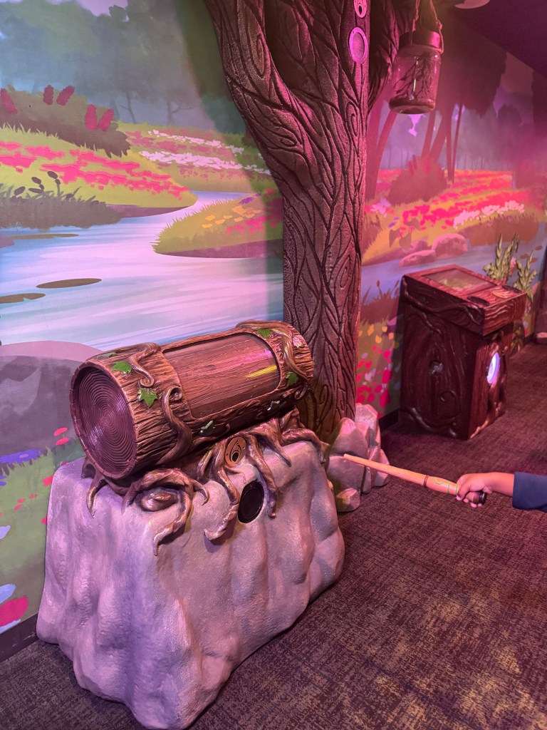 A child using a wand to interact with a magical tree stump and treasure chest in a colorful themed area at Great Wolf Lodge.
