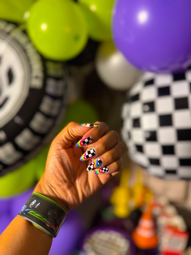 Colorful racing themed nail art with checkered pattern held in front of Monster Jam birthday party decorations and balloons.
