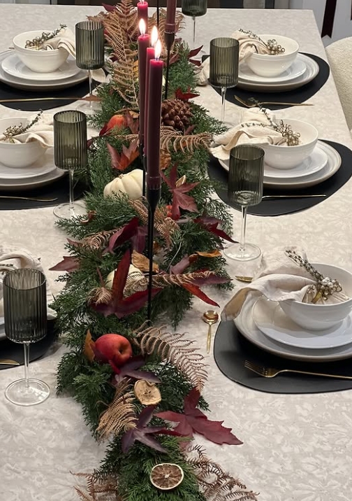 A beautifully styled Thanksgiving table featuring a centerpiece of greenery and autumn leaves, adorned with candles, apple accents, and elegant dinnerware.