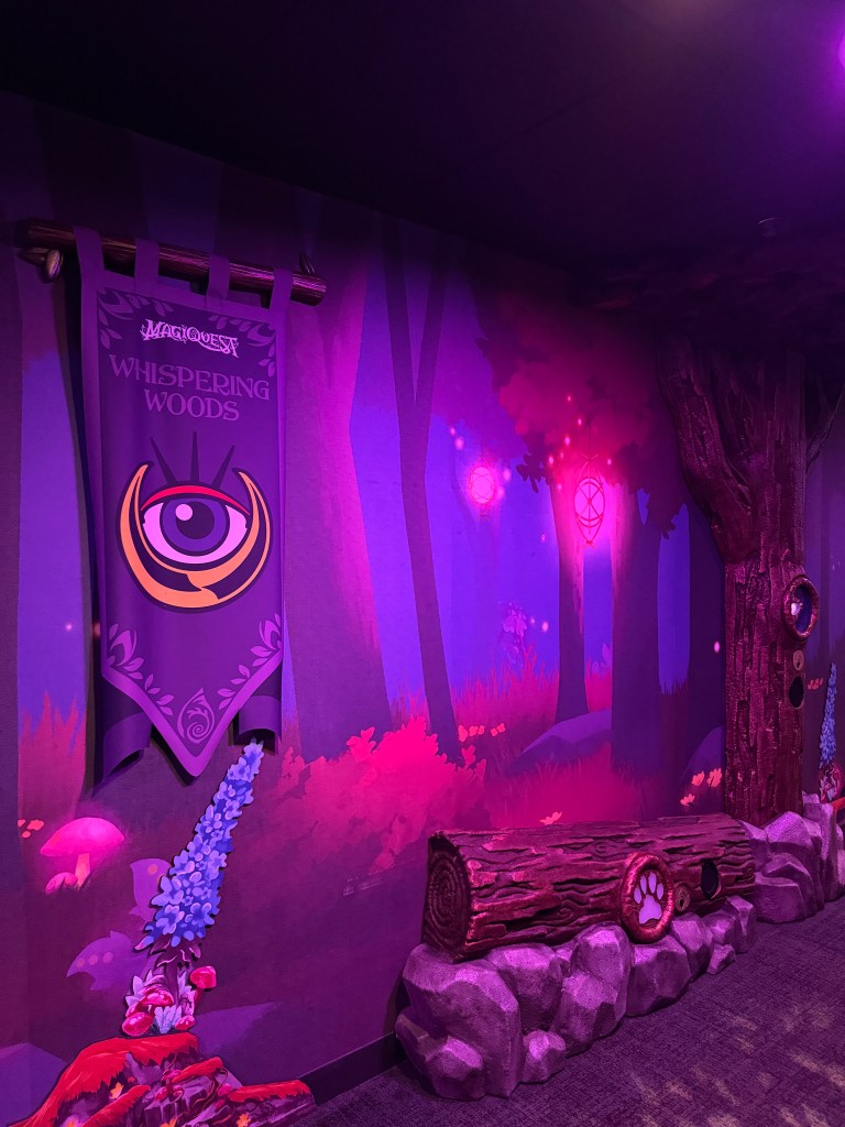 Colorful themed wall featuring the "Whispering Woods" from MagiQuest, with purple and pink hues, showcasing trees, magical elements, and decorative features like a wooden log.