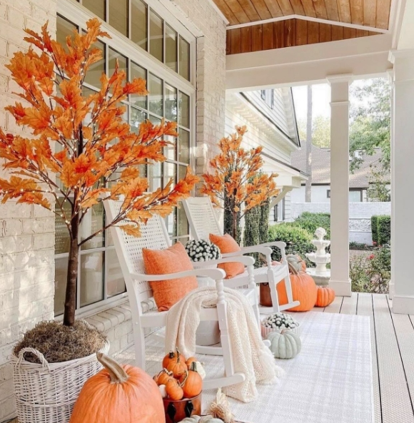 A cozy fall porch featuring orange leaf trees, white rocking chairs with orange pillows, and a variety of pumpkins arranged decoratively.