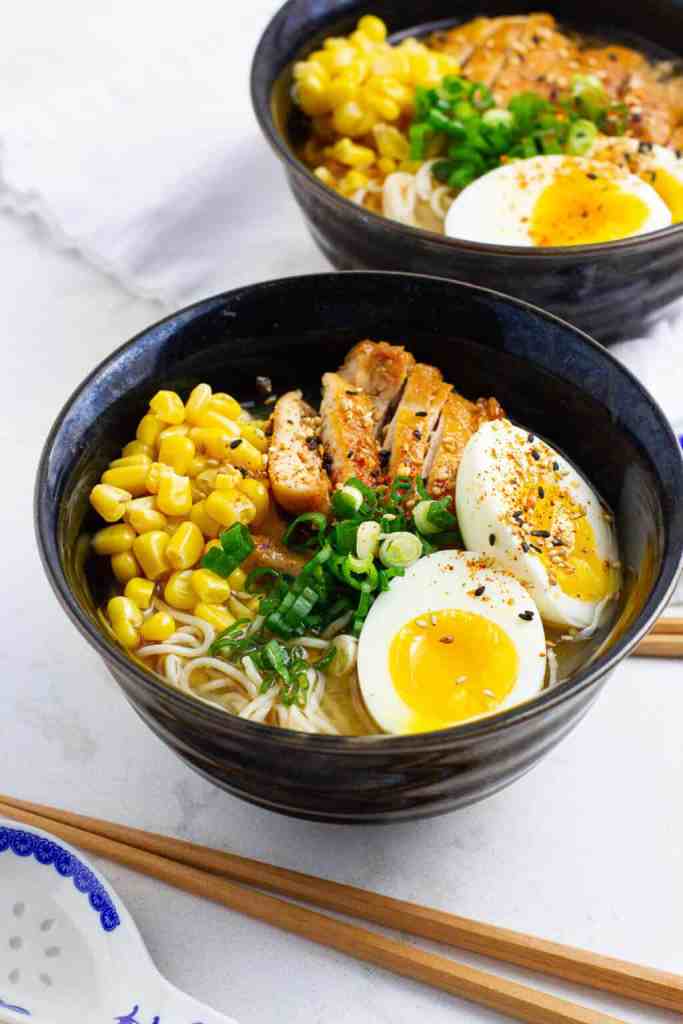 A bowl of chicken ramen featuring sliced grilled chicken, corn, green onions, and a soft-boiled egg on top, served with chopsticks.