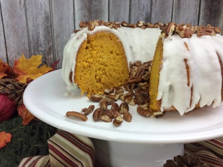 A moist pumpkin Bundt cake topped with white icing, surrounded by chopped pecans on a white cake stand. Autumn leaves and a rustic wooden backdrop add to the fall theme.