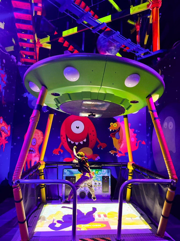 A child playing inside a colorful and vibrant indoor playground featuring a green UFO structure and whimsical monster characters on the walls.