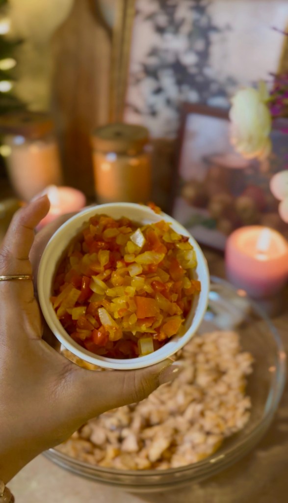A hand holding a small bowl of diced vegetables, including yellow bell peppers and onions, in a kitchen setting with candles and framed pictures in the background.