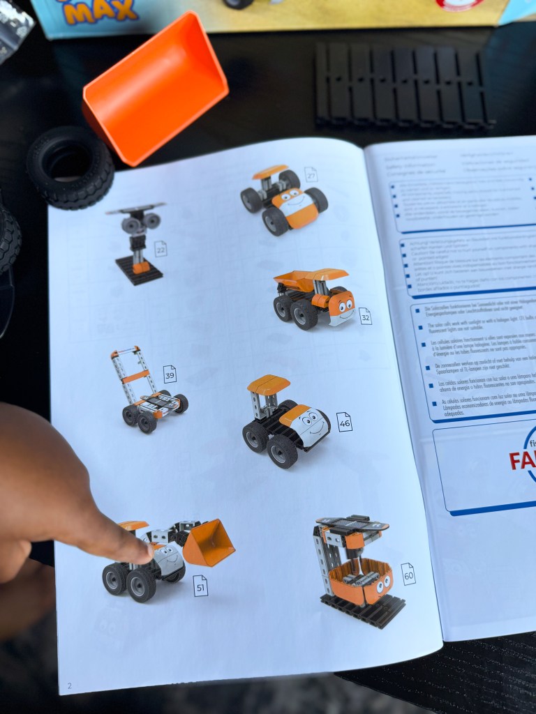 A hand pointing at a page in a construction-themed instruction booklet displaying various colorful toy vehicles, including a dump truck and a loader, with numbered designs and instructions.