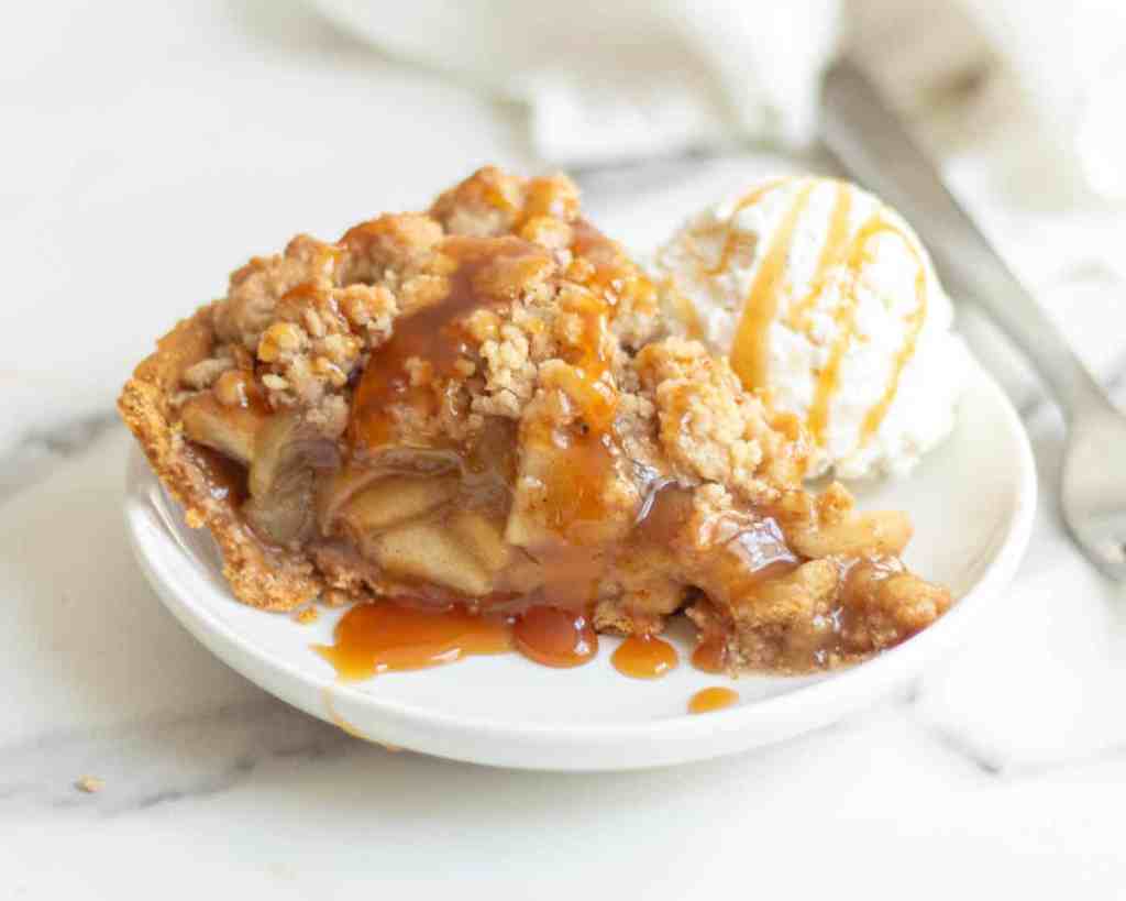 A slice of apple crumble pie served on a white plate, topped with a scoop of vanilla ice cream and drizzled with caramel sauce.