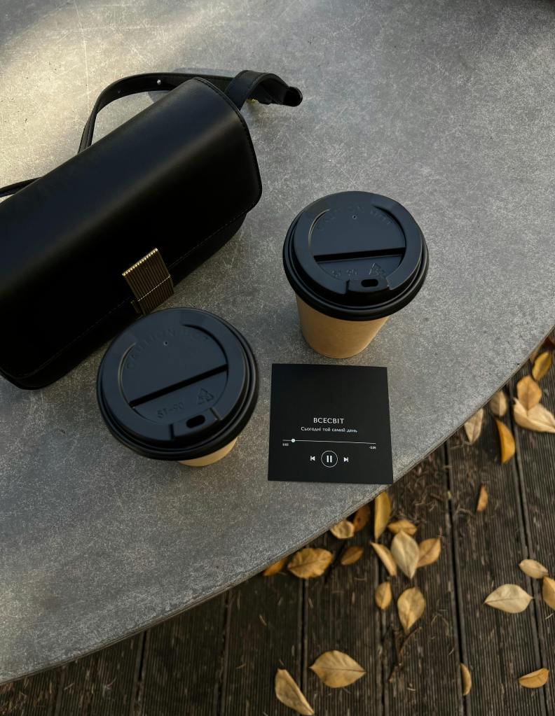 A cozy autumn scene featuring two takeout coffee cups on a table next to a black bag and music playback card, with dried leaves scattered around.