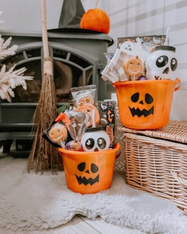 Two orange jack-o'-lantern buckets filled with Halloween treats, including cookies and toys, placed beside a wicker basket in a cozy interior setting.