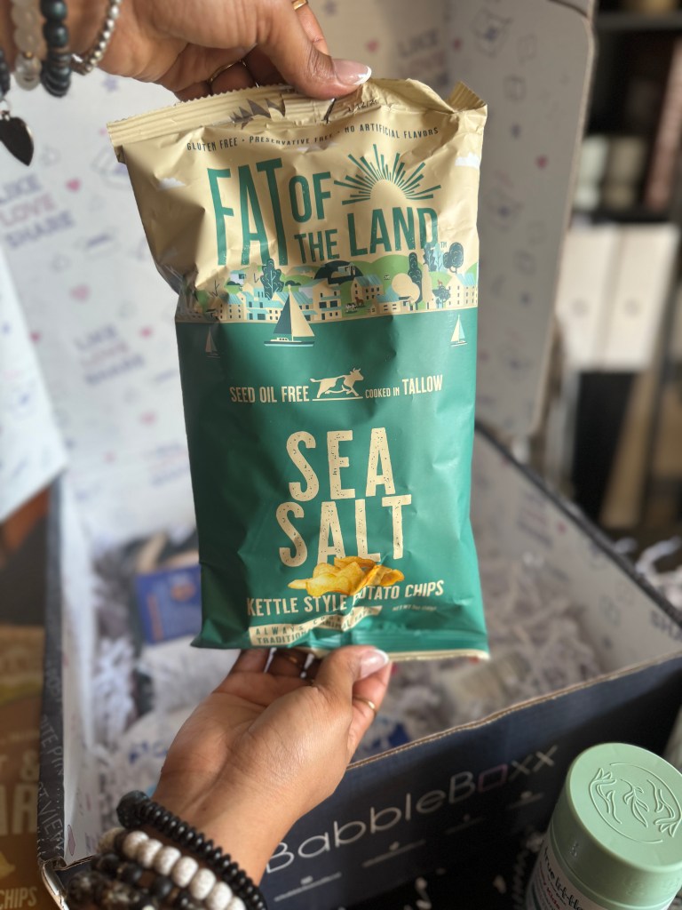 A hand holding a bag of 'Fat of the Land' sea salt kettle style potato chips in front of a BabbleBoxx filled with packing material.