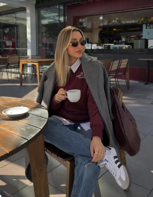 A woman sitting at a café table, wearing a gray coat over a burgundy sweater with a white shirt underneath, holding a cup of coffee. She has on sunglasses and casual sneakers, with jeans and a brown bag placed beside her.