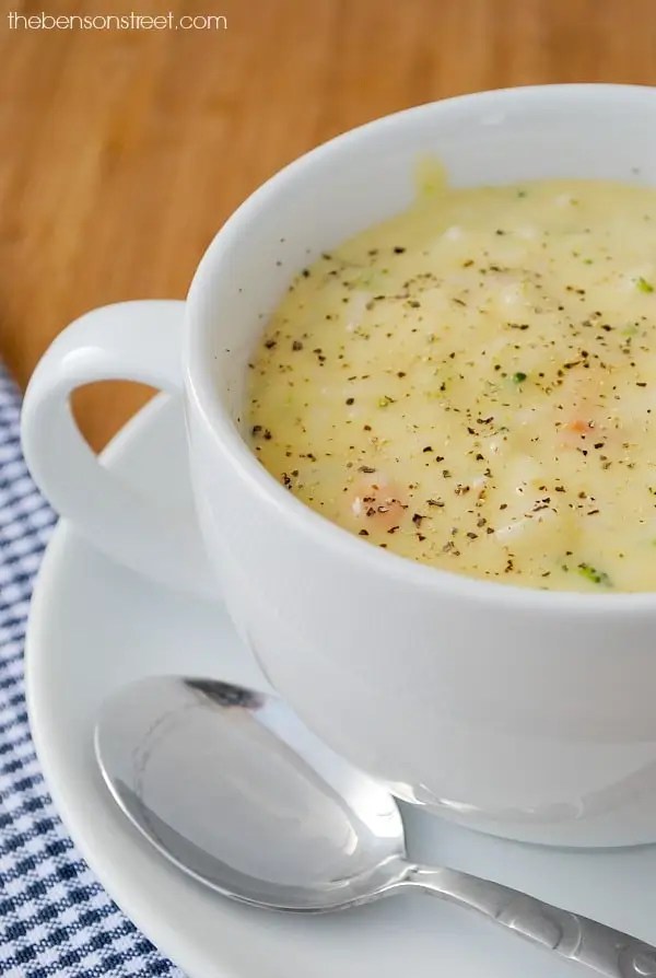 A steaming bowl of creamy soup served in a white cup, garnished with black pepper, resting on a small plate with a spoon beside it.