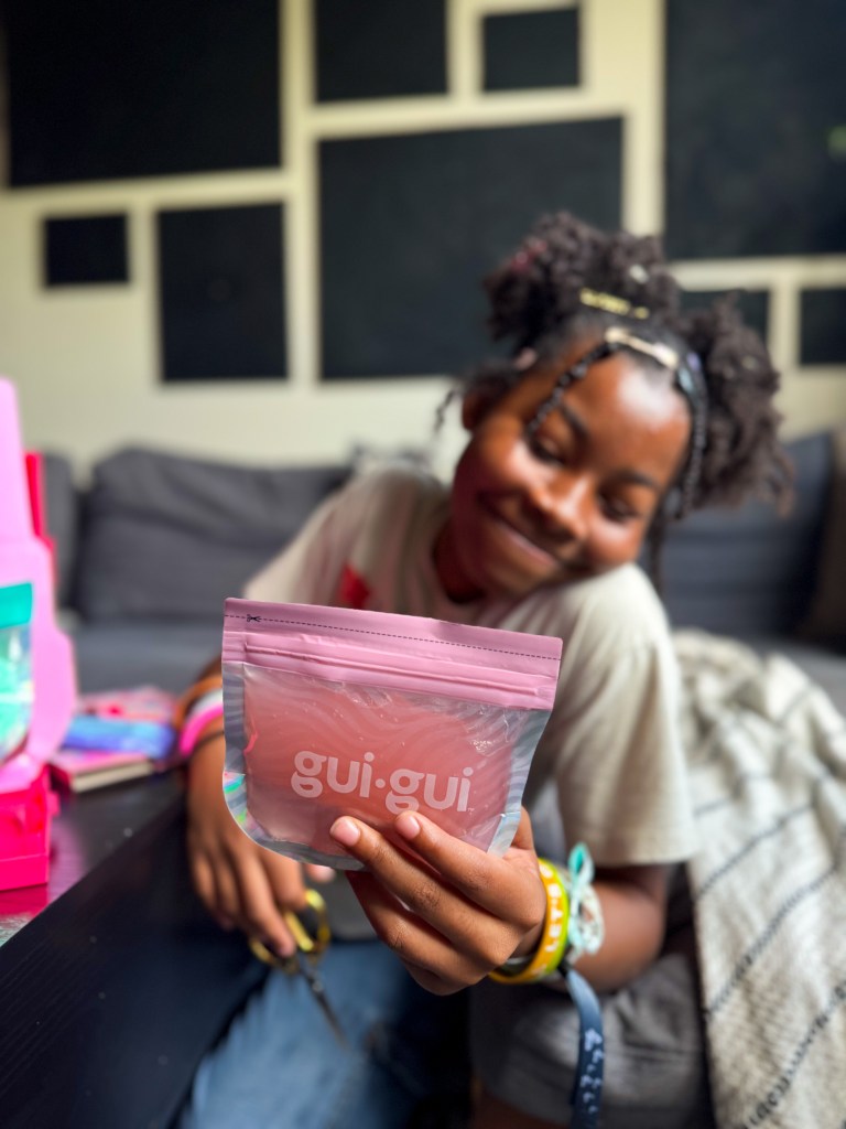 A young person holding a pink pouch labeled 'gui.gui,' sitting on a couch with various colorful items surrounding them.