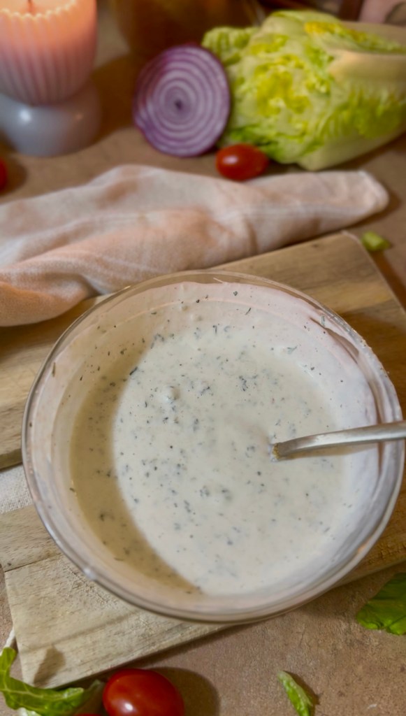 A bowl of creamy ranch dressing with herbs, surrounded by fresh vegetables including lettuce, cherry tomatoes, and red onion on a wooden cutting board.