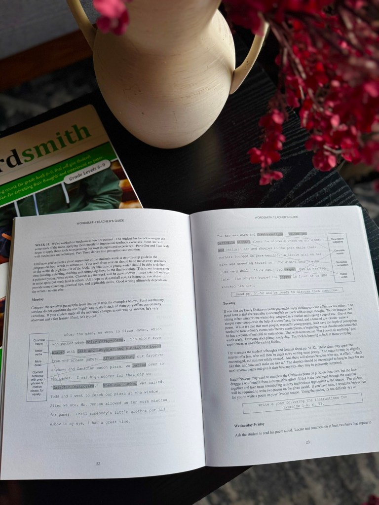 An open 'Wordsmith Teacher's Guide' book on a black table, displaying lesson plans and instructional text. Nearby is a decorative pitcher and a bunch of flowers. A partially visible green book titled 'Wordsmith' is also on the table.