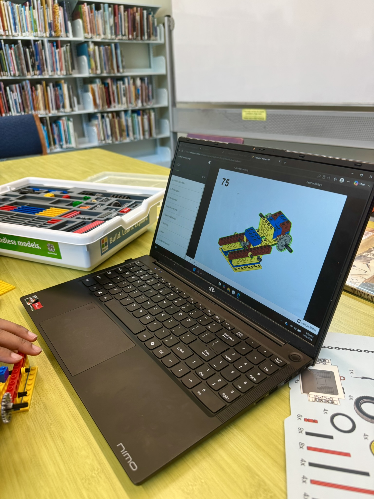 Laptop displaying digital instructions for building a Lego model next to a Lego kit on a table in a library